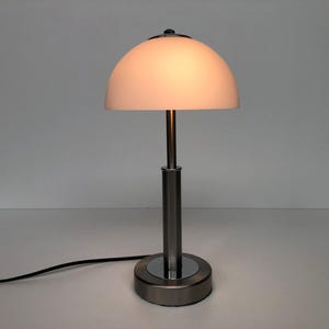 May include: A modern desk lamp with a brushed metal finish. The lamp features a half-dome shade in a soft, warm color, supported by a slender, cylindrical stem. The base is circular, and a black power cord extends from the base.