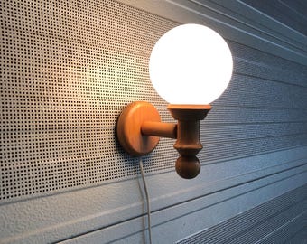 May include: A wooden wall sconce with a round, white globe light. The sconce is made of light-colored wood and has a simple, elegant design. The light is turned on, illuminating the surrounding area.