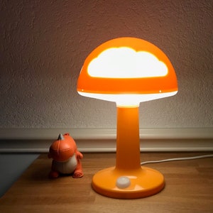 May include: Orange mushroom-shaped table lamp with a white cloud cutout in the shade. The lamp is turned on and illuminating the room.