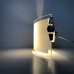 May include: A white curved glass wall sconce with a black switch and cord. The light is on and illuminating the wall.