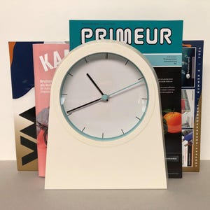 May include: A white clock with a light blue face and black hands. The clock is sitting on a stack of magazines, including one with the word "PRIMEUR" printed on the cover.