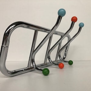 Pode incluir: Um cabide de metal cromado com quatro ganchos curvos. Os ganchos têm extremidades em forma de bola coloridas em azul, laranja e verde. O cabide tem uma estrutura retangular e é projetado para montagem na parede.