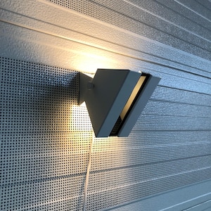 Könnte beinhalten: Eine moderne, graue, geometrische Wandleuchte. Die Leuchte hat eine dreieckige Form mit einer Lichtquelle, die ein warmes Leuchten ausstrahlt. Das Licht ist an einer strukturierten, perforierten Metallwand montiert.