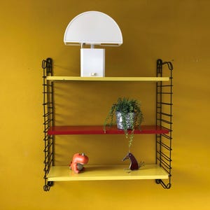May include: A wall-mounted shelving unit with three shelves in yellow and red. A white lamp sits on the top shelf. The shelves hold a potted plant, a small orange dinosaur figurine, and a metal bird sculpture. The unit has a black metal frame.