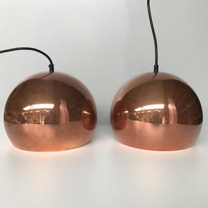 Op de afbeelding: Twee koperen hanglampen met een glanzende, reflecterende afwerking. De lampen zijn bolvormig en hangen aan zwarte snoeren.