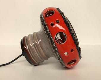Może przedstawiać: Ceramiczna lampa stołowa z czerwonym, błyszczącym kloszem z okrągłymi wycięciami. Klosz jest obramowany teksturowanym czarnym wzorem. Podstawa lampy jest jasnoszara z brązowymi akcentami i czarnym przewodem zasilającym.