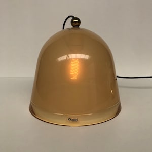 May include: A dome-shaped table lamp with a warm amber hue. The lamp features a gold-colored top and a visible lightbulb inside. The base of the lamp has the text "iGuzzini". A black cord extends from the top.
