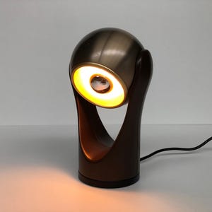 Puede incluir: Una lámpara de escritorio esférica de color bronce con una luz amarilla brillante que emana de la parte delantera. La lámpara tiene una base marrón oscuro y una estructura de soporte curvada. Un cable de alimentación negro se extiende desde la base.
