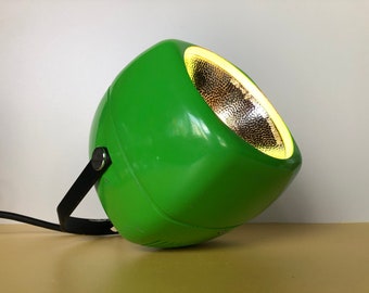 May include: A green spotlight with a black handle and a silver reflector. The light is on and casting a warm glow.