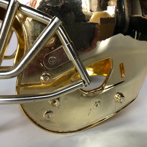 May include: A gold football helmet with a silver face mask. The helmet has a shiny, metallic finish and is decorated with several small screws.