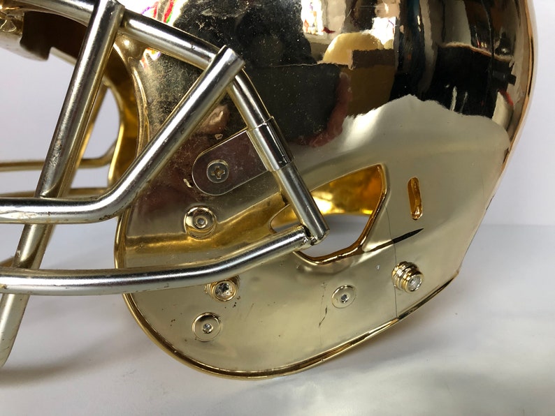 May include: A gold football helmet with a silver face mask. The helmet has a shiny, metallic finish and is decorated with several small screws.