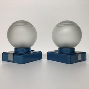 May include: Two matching art deco style lamps. Each lamp features a frosted glass globe with horizontal ridges, atop a square, teal-colored base. A silver metal clip secures the globe to the base. The lamps are displayed on a white surface.