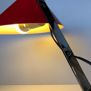 May include: A red and black desk lamp with a conical shade. The lamp is on and the light is shining.