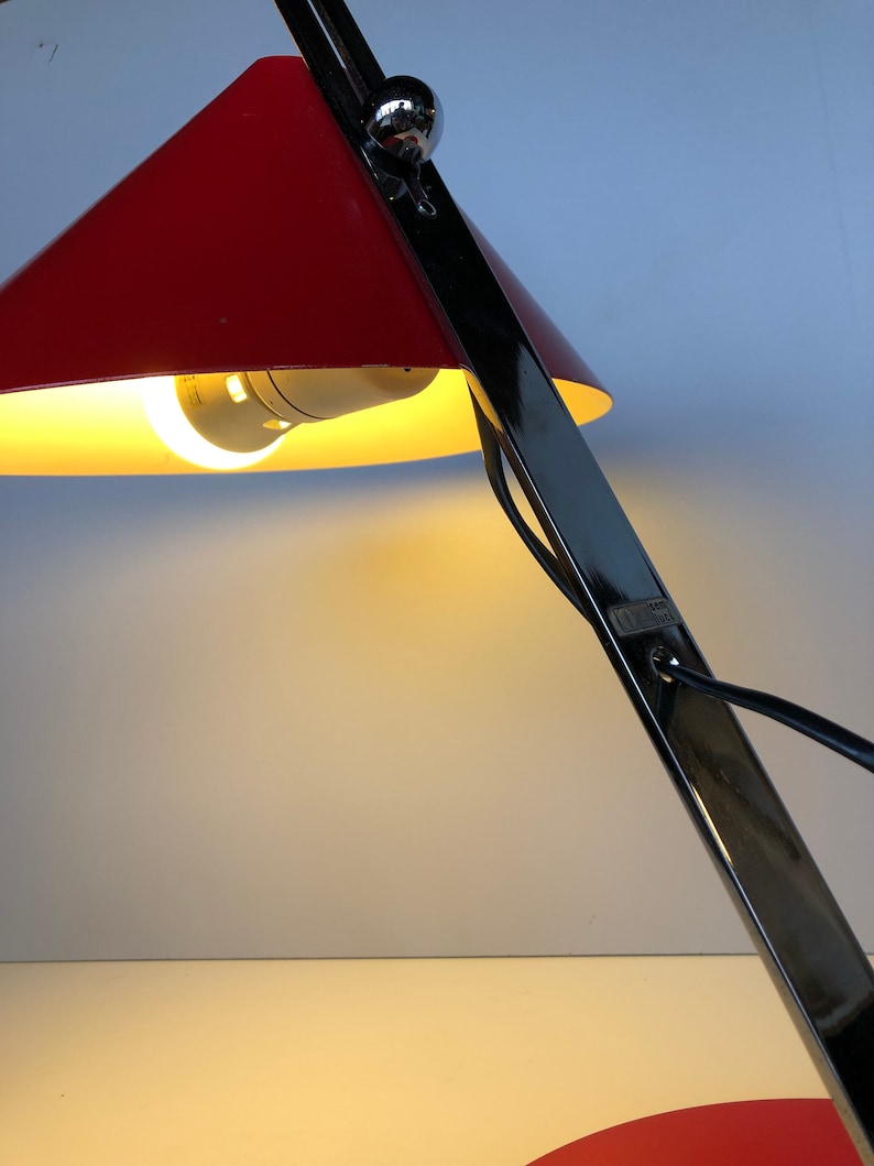 May include: A red and black desk lamp with a conical shade. The lamp is on and the light is shining.