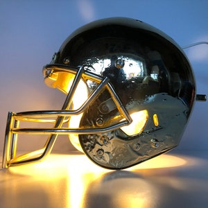 May include: A gold football helmet lamp with a clear light bulb inside. The helmet has a face mask and is lit up.