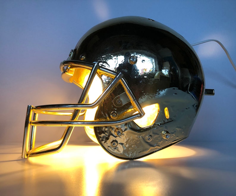 May include: A gold football helmet lamp with a clear light bulb inside. The helmet has a face mask and is lit up.