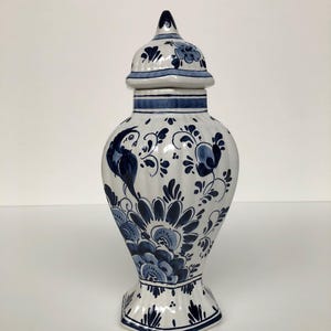 Puede incluir: Un jarrón de cerámica blanca con tapa, adornado con motivos florales y de pájaros azules. El jarrón tiene un cuerpo bulboso y una base hexagonal. La tapa está rematada con un pequeño pináculo. El jarrón mide unos 30 cm de alto.
