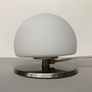 May include: A modern table lamp with a frosted white, dome-shaped shade. The lamp sits on a circular, brushed metal base. A clear cord extends from the base. The lamp provides soft, ambient lighting.