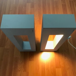 May include: Two gray rectangular lamps with a rectangular cutout in the front. The lamp on the right is lit, casting a warm glow on the wooden floor.