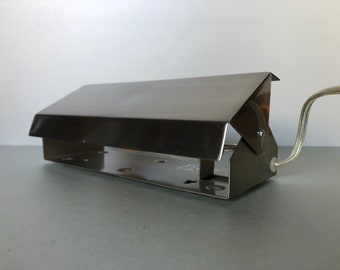 May include: A rectangular, metallic silver-toned wall-mounted picture light fixture with a hinged, adjustable shade. The lamp features a flat base with pre-drilled mounting holes and a clear electrical cord extending from the side of the housing.