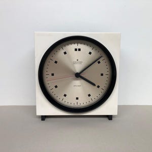 May include: A square white table clock with a circular black frame and a metallic silver face. The clock features black square hour markers, black hour and minute hands, and a thin red second hand. The text Junghans Electronic Ato-Mat is visible.