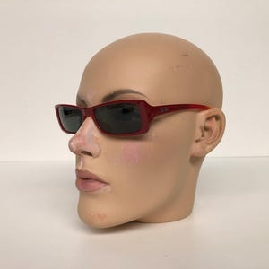 Peut inclure: Lunettes de soleil rectangulaires rouges avec des verres foncés présentées sur une tête de mannequin. Les lunettes de soleil ont une finition brillante et un logo discret sur la branche. La tête de mannequin est de couleur neutre.
