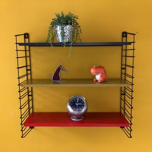 May include: A wall-mounted shelving unit with two black metal side frames and three horizontal shelves in black, gold, and red. The top shelf holds a potted green plant, the middle shelf features a bird figurine and orange dinosaur, and the bottom shelf holds a round silver clock.