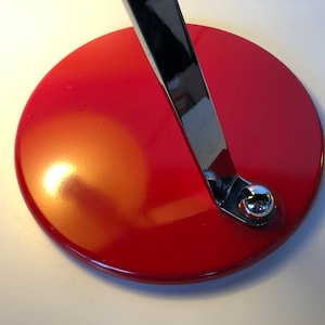 May include: A red circular base with a chrome metal arm attached to it. The base is smooth and shiny.