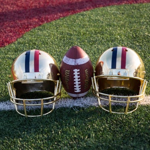 May include: Two gold football helmets with red, white, and blue stripes and a brown leather football with white laces and the word "Wilson" printed on the side. The football is centered between the helmets.