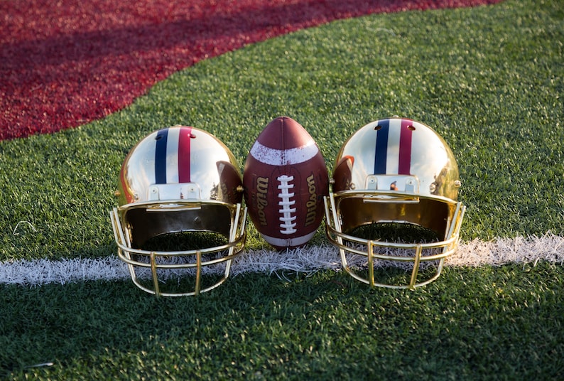 May include: Two gold football helmets with red, white, and blue stripes and a brown leather football with white laces and the word "Wilson" printed on the side. The football is centered between the helmets.