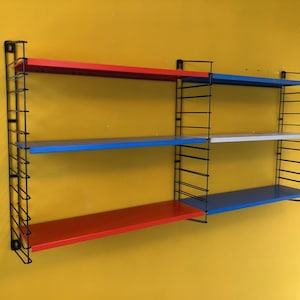 Puede incluir: Estantería de pared con estructura de metal negro y tres estantes. Los estantes están pintados de rojo, azul y blanco. La unidad está montada en una pared amarilla.