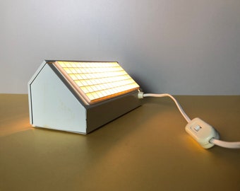 Puede incluir: Una lámpara blanca rectangular inclinada con una cuadrícula de pequeñas casillas iluminadas en la parte superior. La lámpara tiene un cable blanco con un interruptor blanco.