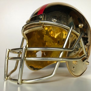 May include: A gold football helmet with a red and blue stripe on the side. The helmet has a face mask and is made of metal.