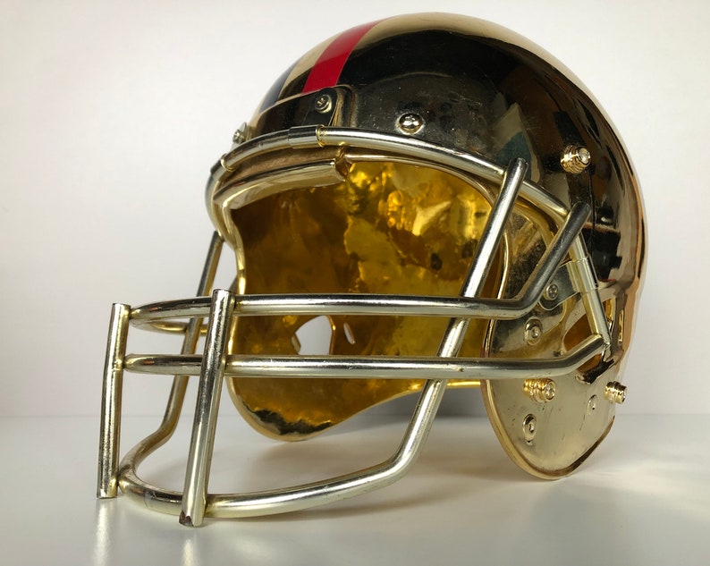 May include: A gold football helmet with a red and blue stripe on the side. The helmet has a face mask and is made of metal.