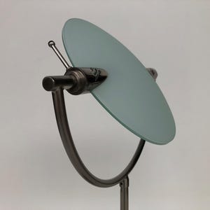 Op de afbeelding: Een moderne make-upspiegel met een cirkelvormige, lichtblauwe glazen spiegel. De spiegel wordt ondersteund door een gebogen, donker metalen frame en een verticale paal. Een klein, dun antenne-achtig detail steekt uit het frame van de spiegel.