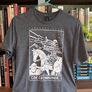 Può includere: T-shirt grigio scuro con un'illustrazione in bianco e nero di uno scheletro che suona il trombone a cavallo. Il testo "THE TROMBONER" è sotto l'immagine. La maglietta è appesa a una gruccia verde.