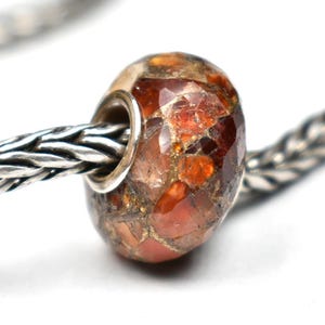 May include: A faceted bead with a mix of orange, brown, and tan tones. The bead is strung on a silver chain. The bead has a metallic silver core and is likely a charm or jewelry component.
