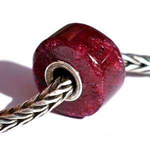 Puede incluir: Una cuenta de piedra preciosa cilíndrica de color rojo intenso con una superficie cristalina texturizada y un núcleo de metal plateado pulido, ensartada en una cadena trenzada de tono plateado. La cuenta presenta inclusiones.