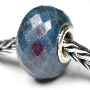 Peut inclure: Une perle facettée bleu-gris avec des éclats rouges, enfilée sur une chaîne argentée. La perle a un cœur argenté métallique et est probablement un breloque ou un composant de bijou. La surface de la perle reflète la lumière, soulignant ses coupes géométriques.