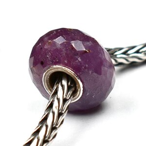 May include: A faceted, deep purple-red gemstone bead with a smooth, polished silver-toned metal core, threaded onto a braided silver-toned chain. The bead has a rounded, slightly flattened shape with multiple flat surfaces reflecting light.