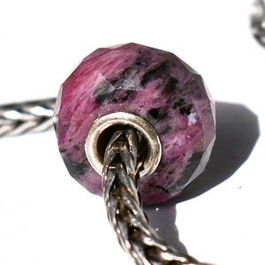 May include: A faceted, round bead made of mottled magenta and black stone, featuring a central silver-toned metal core. The bead is threaded onto a silver-toned chain with a woven, snake-like texture that extends from the center of the bead.