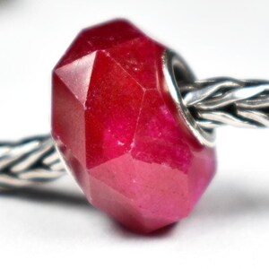May include: A faceted, deep red bead with a metallic silver core. The bead is a geometric shape with multiple flat surfaces, reflecting light. The bead is strung on a silver chain, creating a jewelry component.