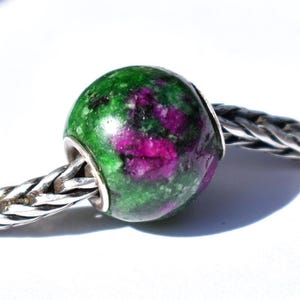 May include: A spherical bead made of green zoisite with inclusions of magenta ruby, featuring a polished surface. The bead is threaded onto a silver chain with a braided, woven metal texture that extends horizontally from both sides of the bead.