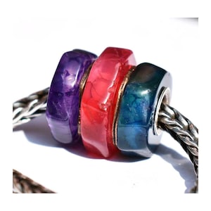 May include: Close-up of three colorful beads on a silver chain. The beads are purple, red, and blue, with a metallic gold accent. The beads are a mix of translucent and opaque materials, creating a unique visual texture.