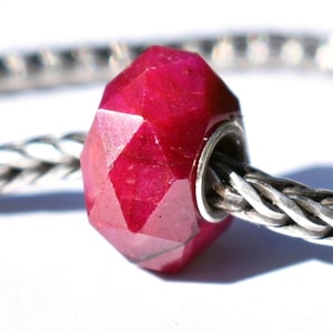 Puede incluir: Una cuenta de piedra preciosa facetada de color rojo intenso con un núcleo de metal plateado. La cuenta está ensartada en una cadena plateada, mostrando su diseño geométrico y su color vibrante. Las superficies pulidas de la cuenta reflejan la luz.