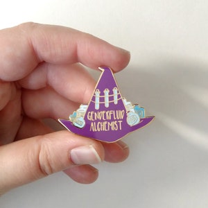 Może przedstawiać: Fioletowa szpilka emaliowana w kształcie kapelusza czarownicy z złotymi akcentami. Kapelusz jest wypełniony butelkami z eliksirami i probówkami. Tekst na szpilce brzmi "Genderfluid Alchemist".