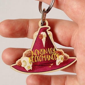 Puede incluir: Un llavero de madera con un diseño de sombrero de bruja burdeos. El sombrero tiene las palabras "NONBINARY NECROMANCER" en letras doradas, con detalles de calaveras y rosas. Está unido a un llavero plateado.