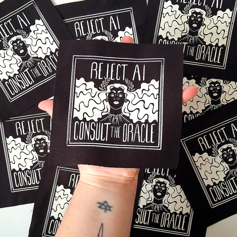 May include: A square black fabric patch featuring a white screen-printed graphic of a person with wavy hair wearing a laurel wreath. The text REJECT AI is printed above the figure, and CONSULT THE ORACLE is printed below, all within a square border.
