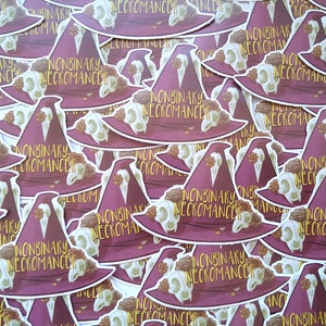 May include: A close-up of a pile of stickers featuring a witch's hat with the text "Nonbinary Necromancer" repeated on each sticker. The stickers are purple and have gold accents.