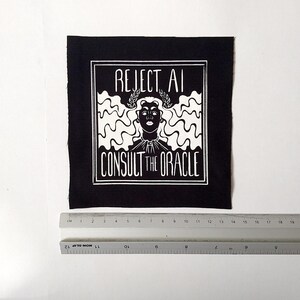 May include: A square black fabric patch featuring a white screen-printed graphic of a person with wavy hair wearing a laurel wreath. The text reads REJECT AI CONSULT THE ORACLE. A ruler placed below shows the patch is approximately 14 cm wide.
